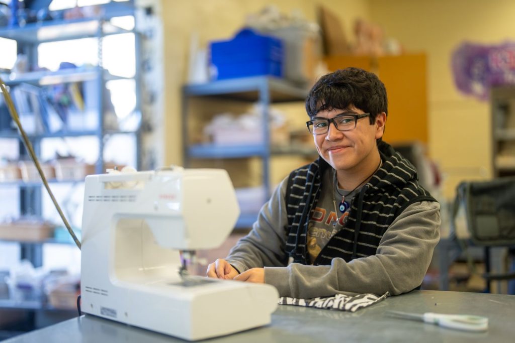 Una estudiante sonríe mientras está sentada frente a una máquina de coser, rodeada de materiales para manualidades en un aula.