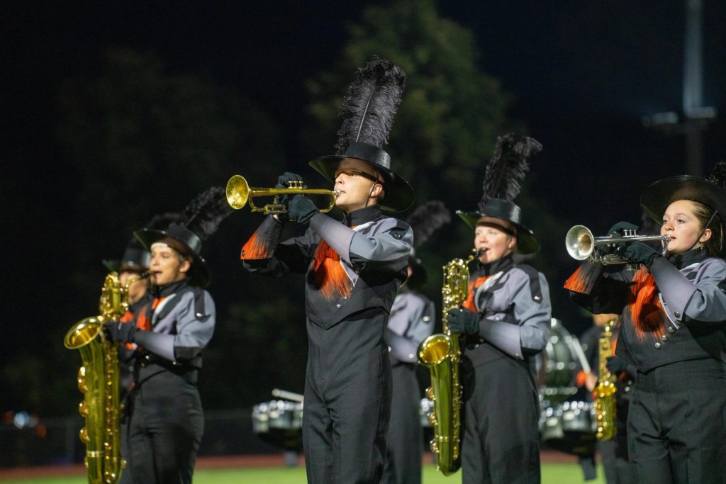 Los miembros de la banda de música, vestidos con sus uniformes, tocan instrumentos de metal bajo las luces del estadio por la noche.