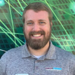 Smiling man with a beard wearing a gray polo shirt stands in front of a green patterned background.