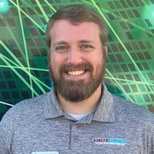 Smiling man with a beard wearing a gray polo shirt stands in front of a green patterned background.