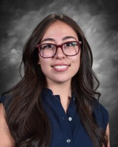A young woman with long dark hair and glasses smiles at the camera, wearing a navy blue blouse against a gray backdrop.