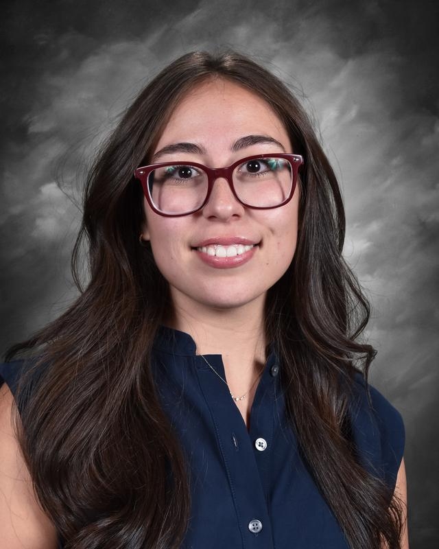 A young woman with long dark hair and glasses smiles at the camera, wearing a navy blue blouse against a gray backdrop.