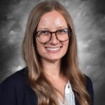 Smiling woman with long brown hair and glasses, wearing a navy cardigan over a polka dot shirt, against a gray background.