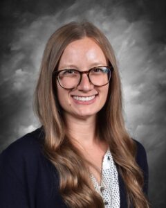 Smiling woman with long brown hair and glasses, wearing a navy cardigan over a polka dot shirt, against a gray background.