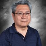 A man with gray hair and glasses smiles, wearing a navy blue polo shirt against a gray background.