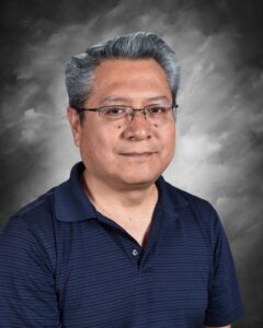 A man with gray hair and glasses smiles, wearing a navy blue polo shirt against a gray background.