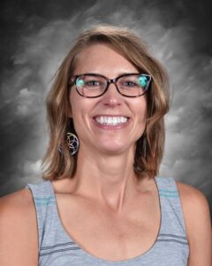 Smiling woman with short brown hair, glasses, and colorful earrings, wearing a gray tank top against a dark background.