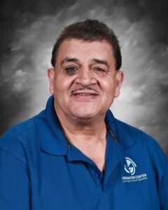 A smiling man with short hair, wearing a blue shirt with a logo, poses for a portrait against a gray background.