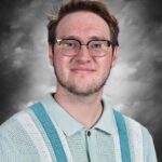 A young man with glasses smiles, wearing a light blue and white striped sweater, against a gray background.