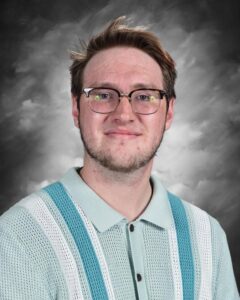 A young man with glasses smiles, wearing a light blue and white striped sweater, against a gray background.