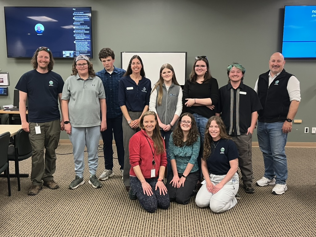 A diverse group of eleven people poses together in a classroom setting, with a screen displaying information behind them.