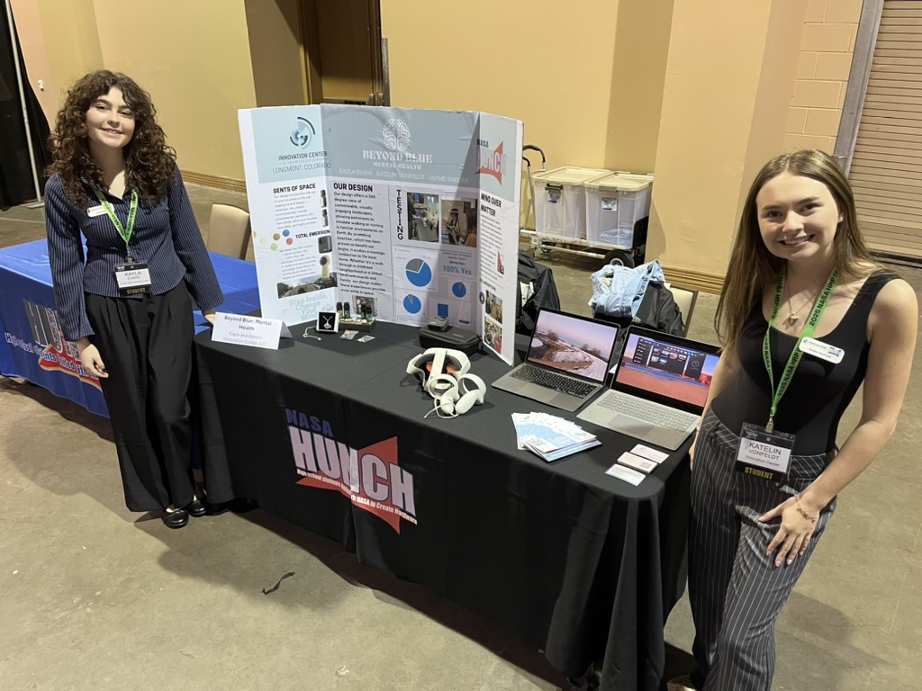 Two young women stand beside a display table featuring project materials, laptops, and promotional items at an event.
