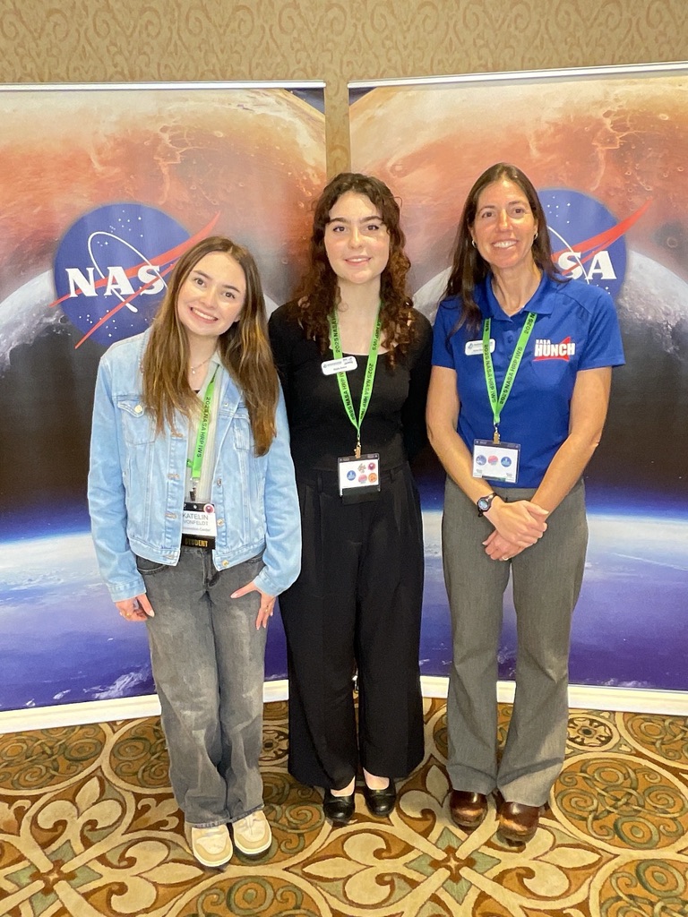 Three women stand together in front of NASA banners, smiling and wearing name tags at an event.