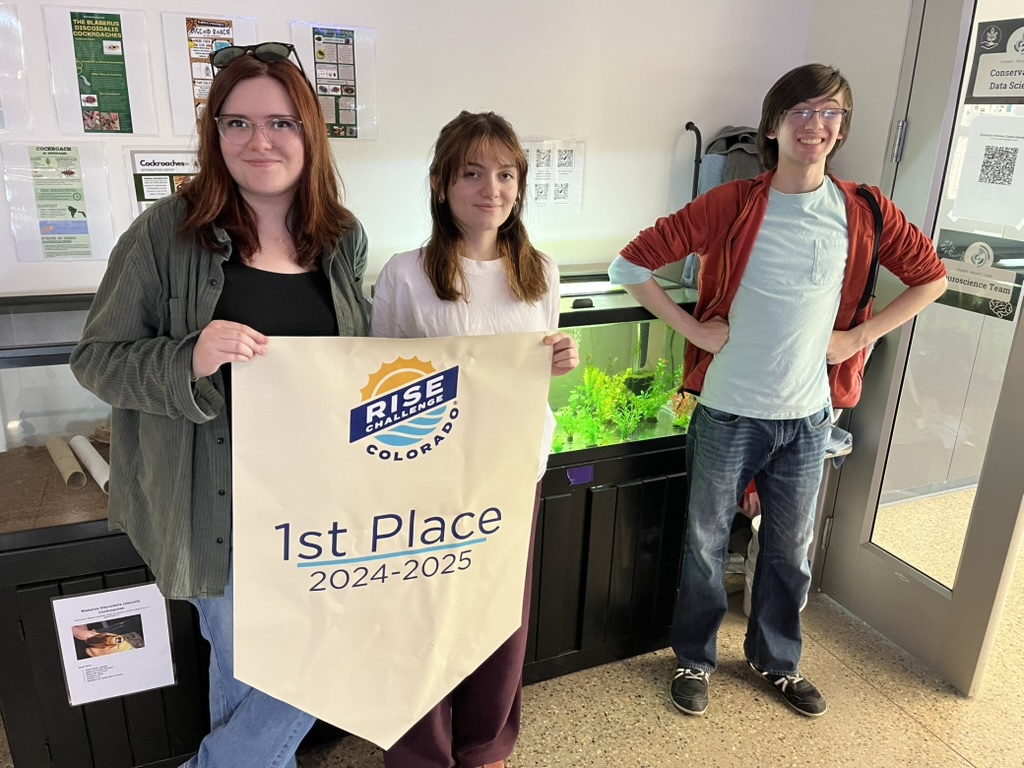 Three students proudly display a "1st Place 2024-2025" banner in front of an aquarium in a school setting.