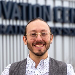 A smiling person with glasses and a beard stands in front of a sign that reads "Innovation Center."
