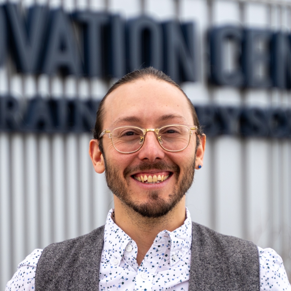 A smiling person with glasses and a beard stands in front of a sign that reads "Innovation Center."