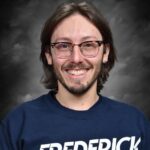 A smiling person with long hair and glasses wears a blue sweatshirt with "Frederick Orchestra" printed on it.