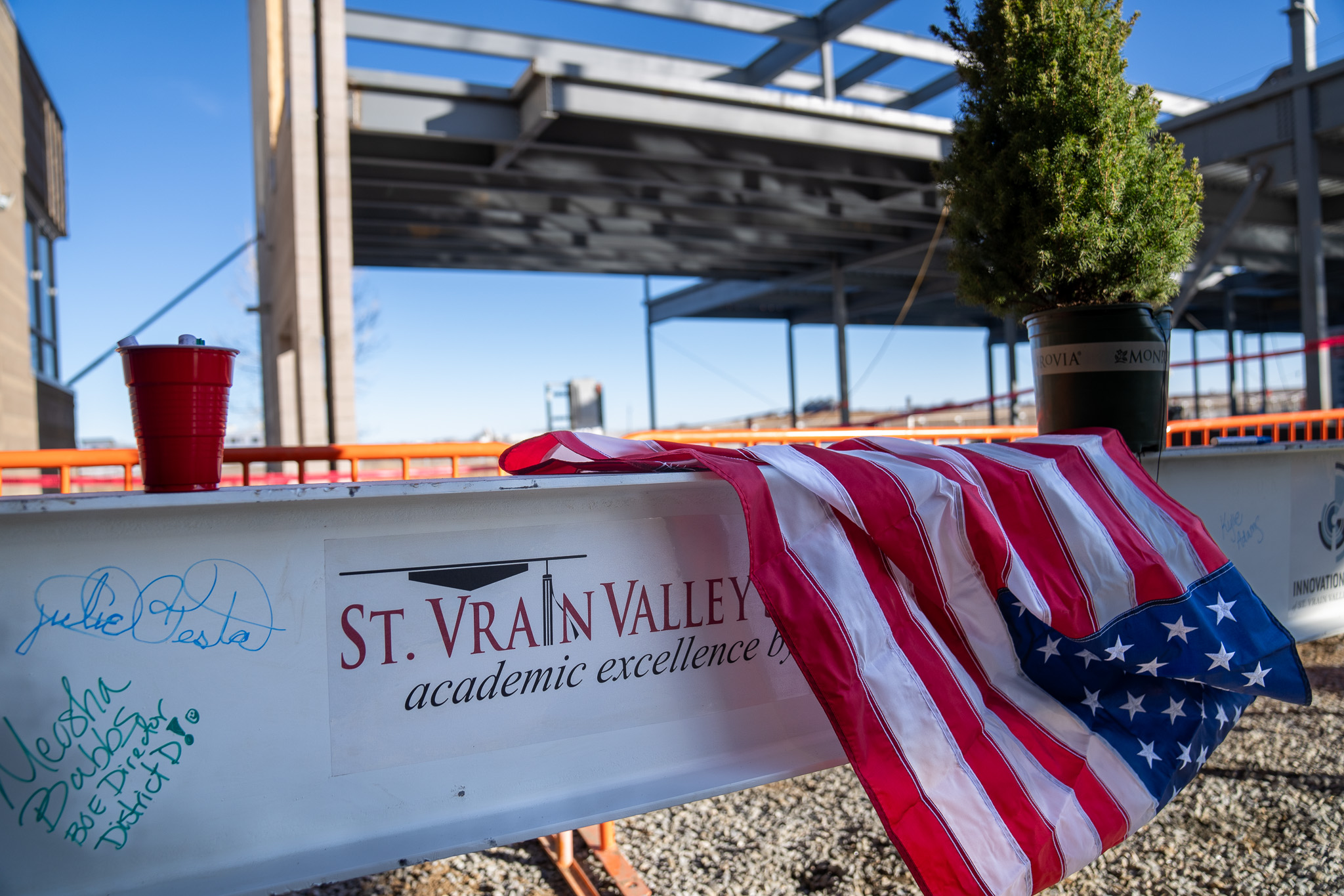 A U.S. flag drapes over a steel beam labeled "St. Vrain Valley," with a red cup and a small potted tree nearby.