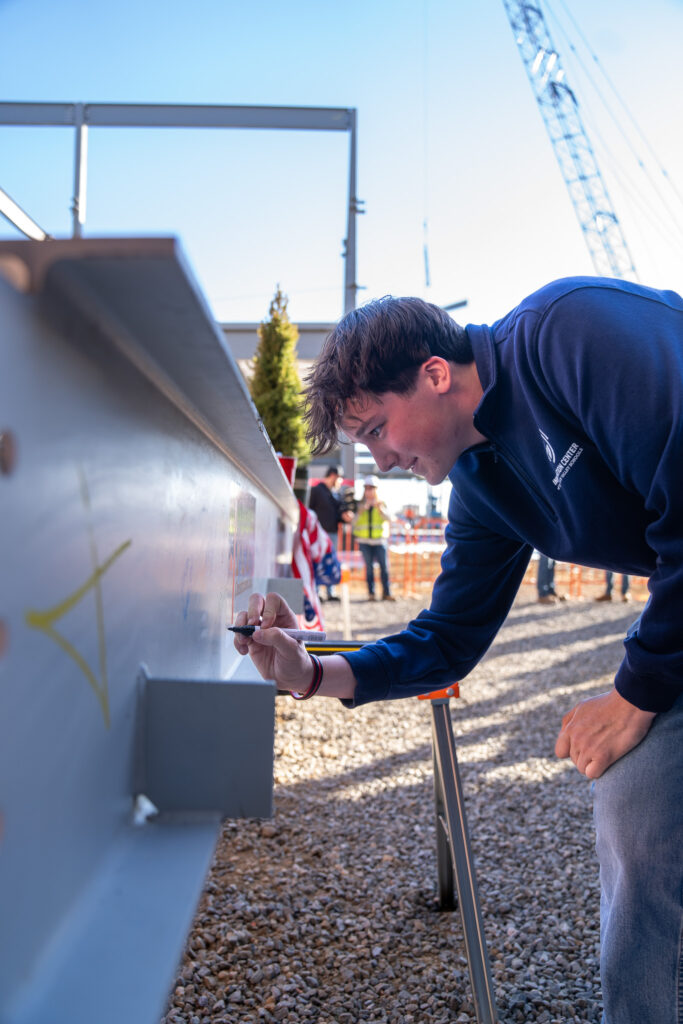 A young man is signing a steel beam at a construction site, with workers and an American flag in the background.
