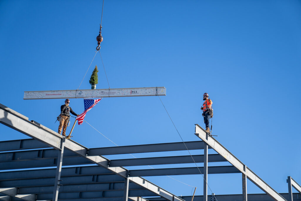 Two construction workers on a steel beam, one holding an American flag, under a clear blue sky.