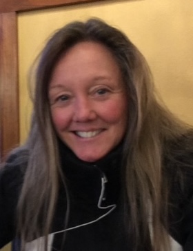 A smiling person with long hair wearing a black and white jacket sits indoors.