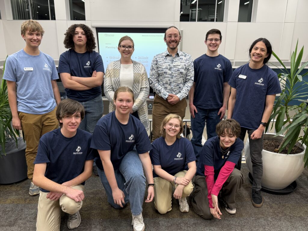 A group of nine people, including students and an adult, pose together in a modern indoor setting with plants.