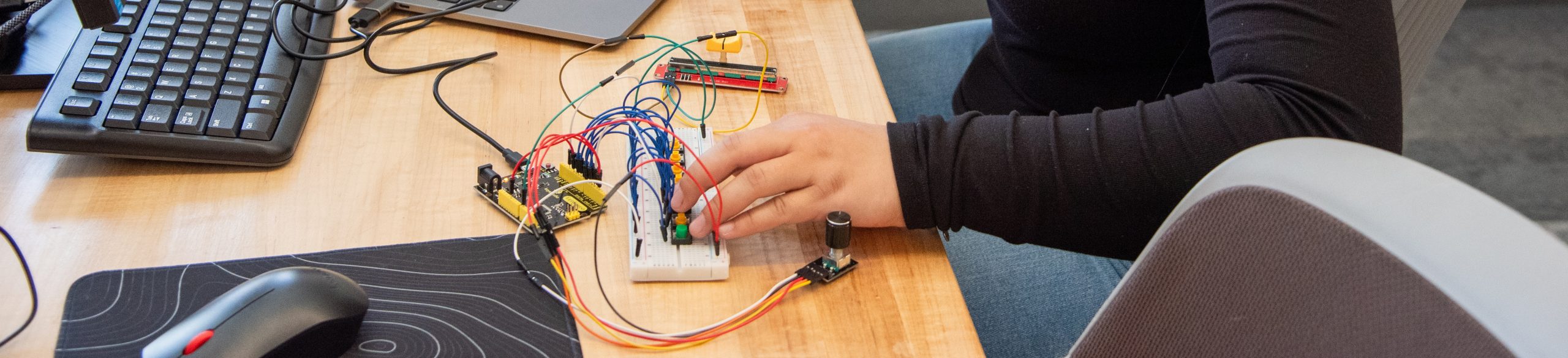 Una persona conecta cables en una placa de pruebas, con un teclado y un ratón cerca, mientras trabaja en un proyecto de electrónica.
