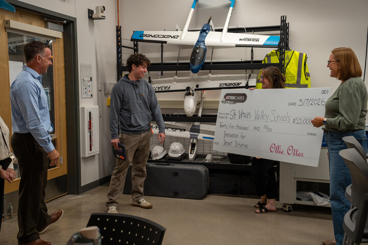 A group of people stand in a room with drones, holding a large check for St. Vrain Valley Schools.