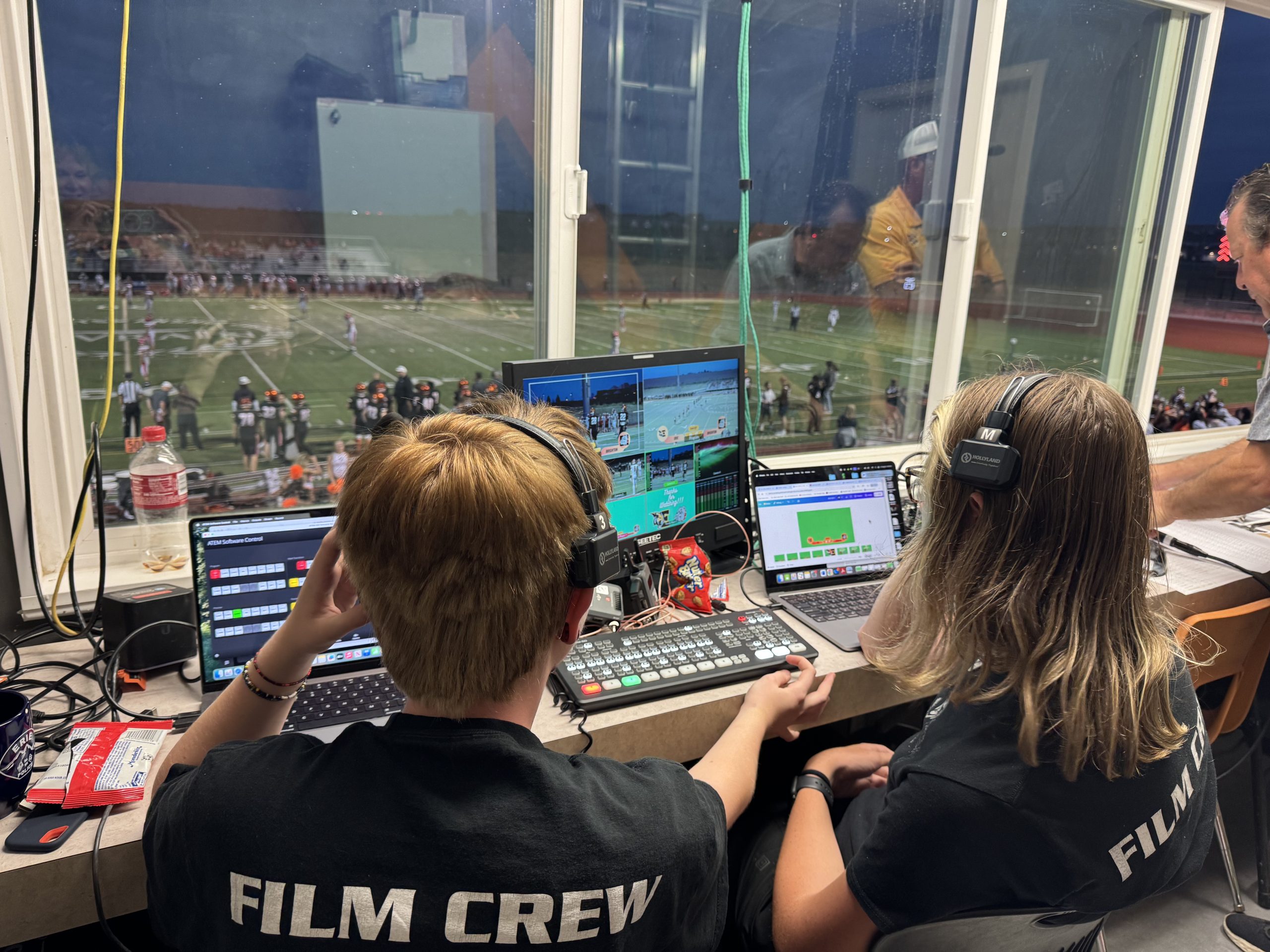 Dos miembros del equipo de filmación manejan el equipo de transmisión en vivo desde una cabina con vista a un partido de fútbol.