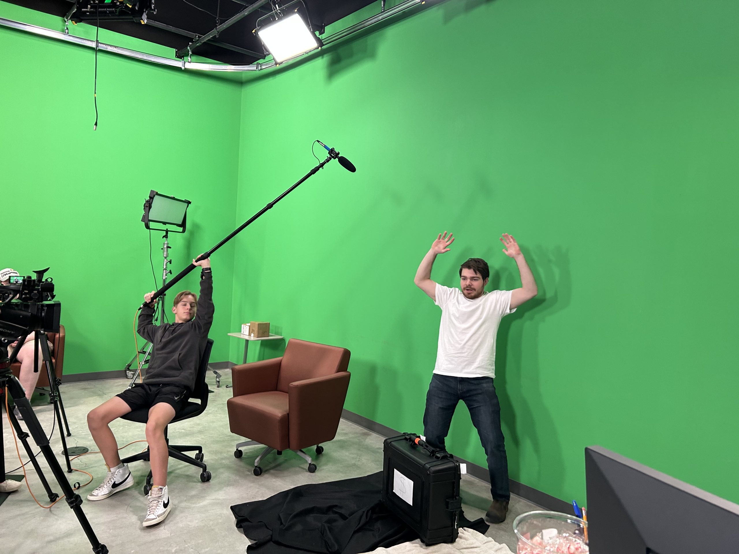 Two people in a green screen room, one holding a boom mic, the other posing with raised arms.